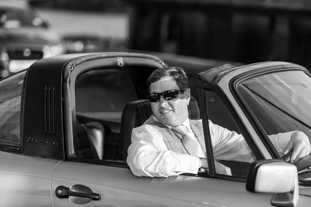 Man wearing sunglasses, dress shirt, tie, and vest, sitting in the driver's seat of a vintage Porsche Targa convertible with the door open.