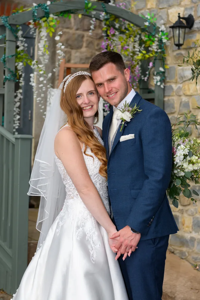 Smiling bride in a white gown and veil standing with her groom in a blue suit under a floral archway.