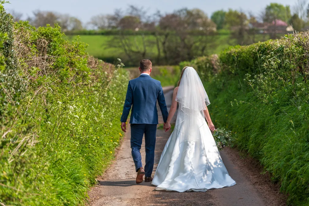 Bride and groom holding hands walking down a country lane bordered by green hedges.