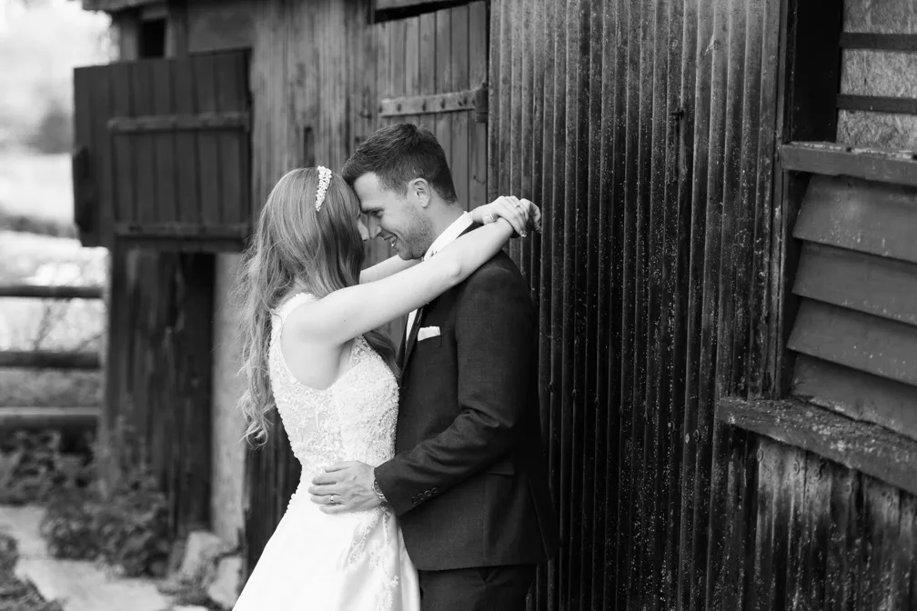 Bride and groom embracing closely with foreheads touching in front of a rustic wooden wall.