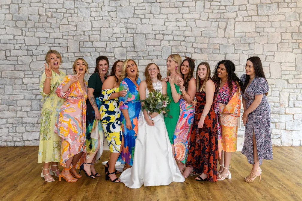 Bride in white dress holding bouquet surrounded by ten women in colorful dresses smiling and posing indoors against a stone wall.