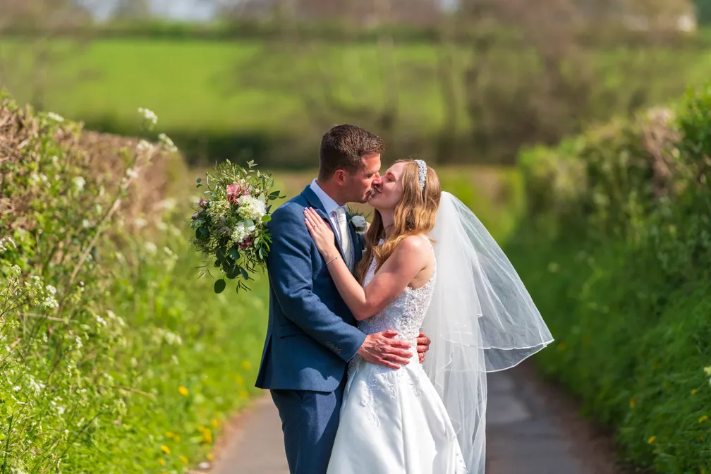 Bride and groom in wedding attire kissing on a country path surrounded by green hedges and fields.