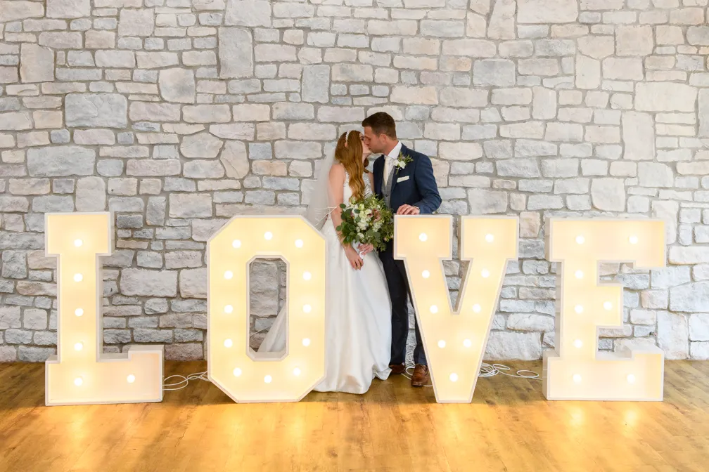 Bride and groom kissing behind illuminated LOVE letters on wooden floor with stone wall background.