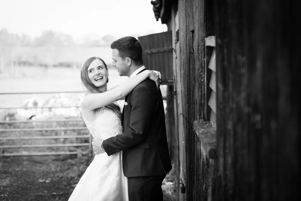 Bride and groom embracing and smiling outdoors near a wooden fence in a black and white wedding photo.