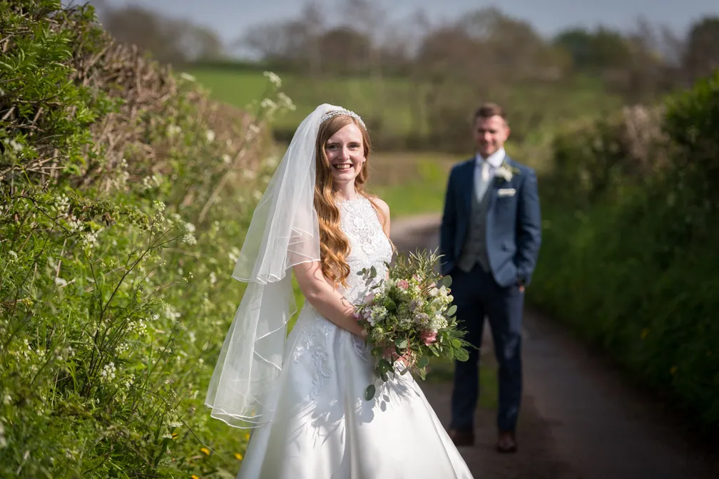 Smiling bride in white wedding dress holding a bouquet, with groom blurred in the background on a country path.