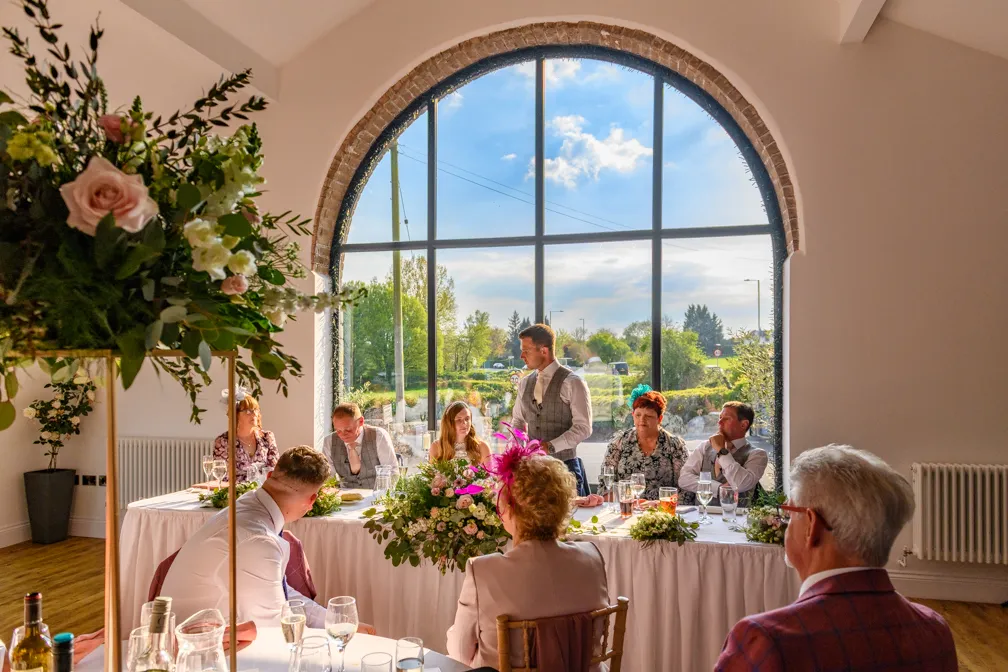 Wedding reception with groom standing at a decorated head table in front of a large arched window overlooking greenery.