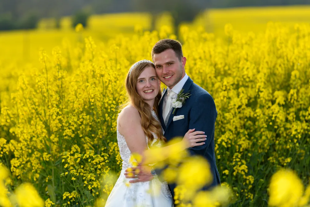 Bride and groom embracing in a field of yellow flowers during sunset.