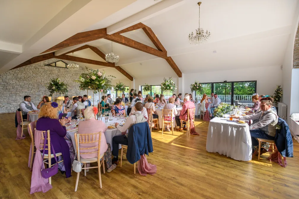 Wedding reception in a bright hall with wooden ceiling beams, decorated tables, and guests seated around enjoying the event.
