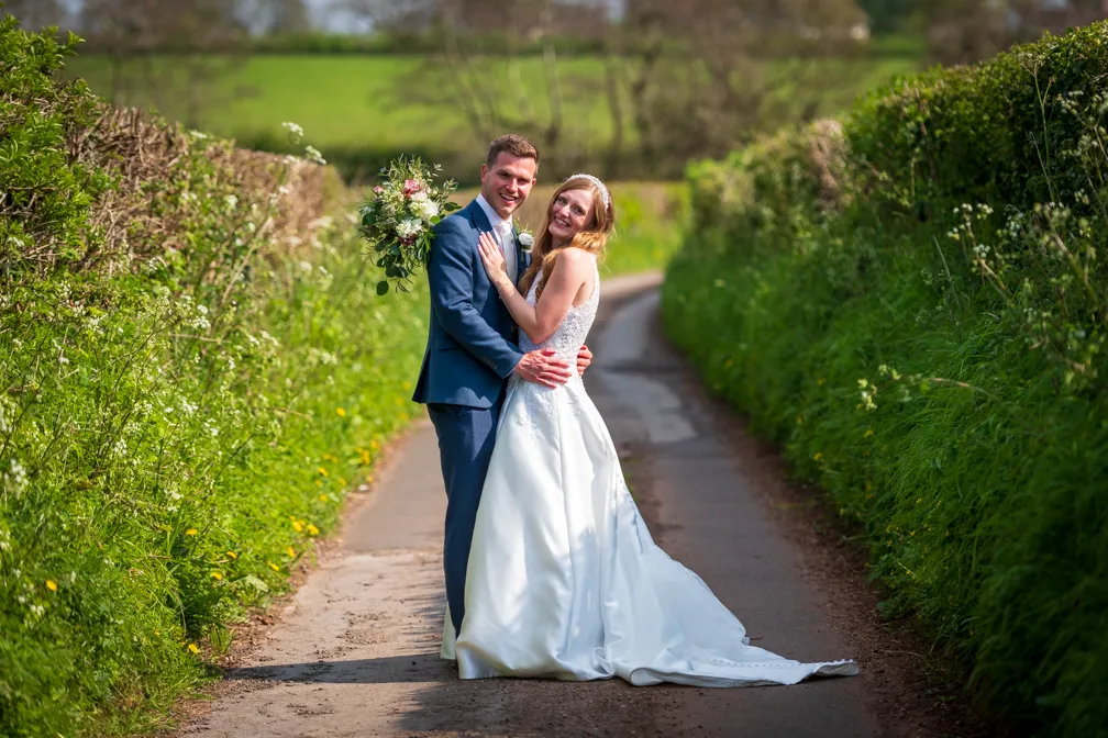Bride and groom embracing on a countryside lane lined with green hedges during their wedding.
