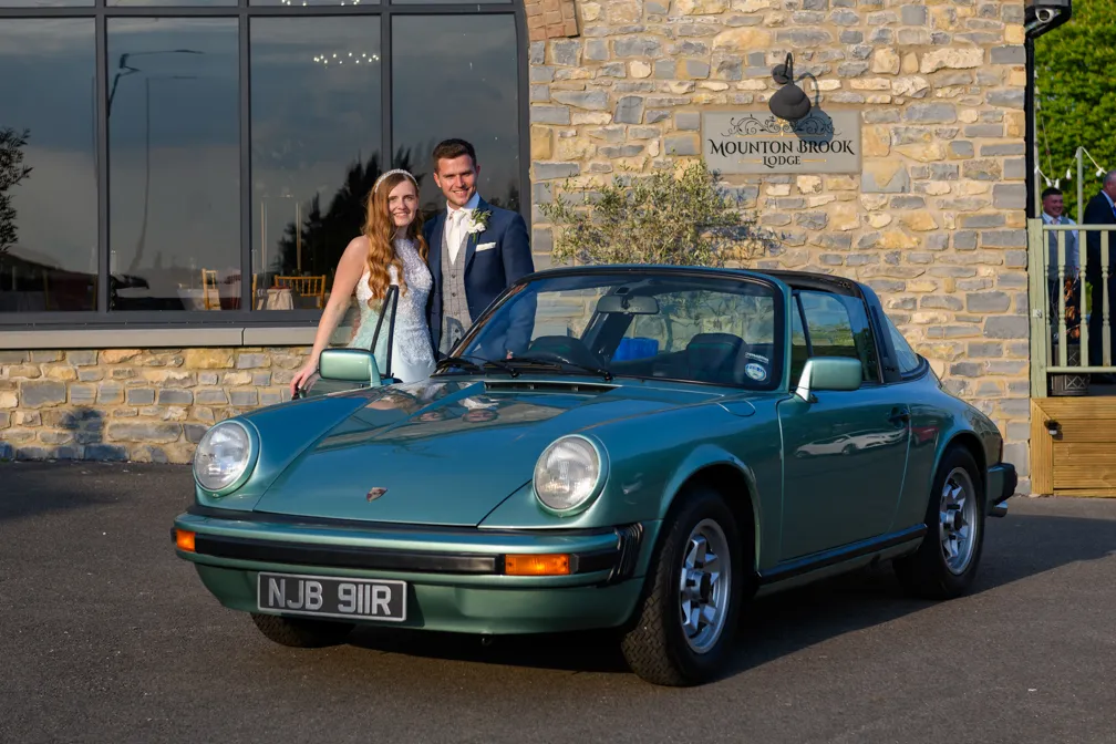 Bride and groom posing beside a green classic Porsche Targa outside Mounton Brook Lodge on a sunny day.