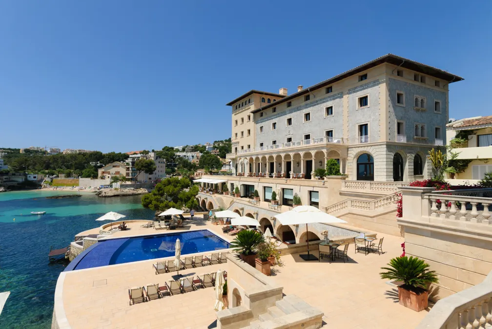 Elegant coastal hotel with a terrace featuring an infinity pool, lounge chairs, umbrellas, and sea views.
