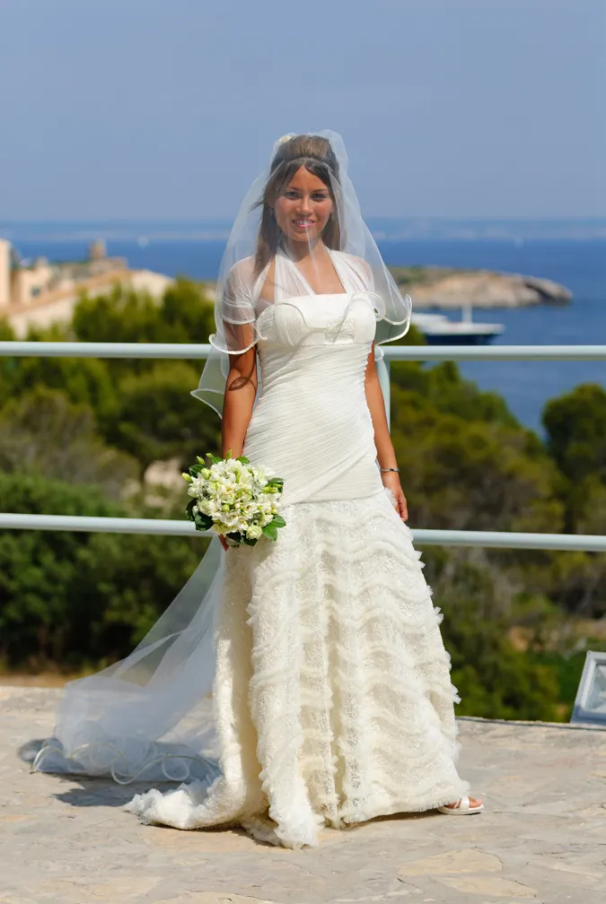 Smiling bride in a white wedding dress and veil holding a bouquet with a seaside background.