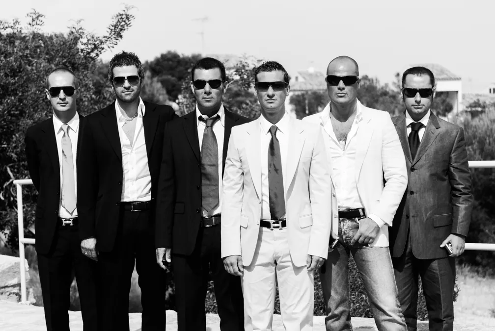 Group of six men standing outdoors in suits and sunglasses, three wearing lighter suits and three in darker suits.