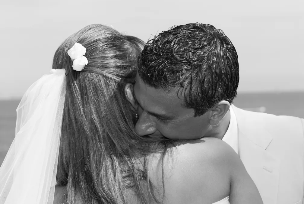 Bride and groom embracing closely outdoors near water, the groom gently kissing the bride's shoulder.