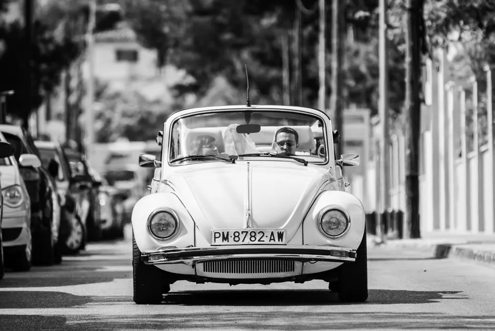 Front view of a white vintage convertible car with two passengers driving on a street lined with parked cars and trees.