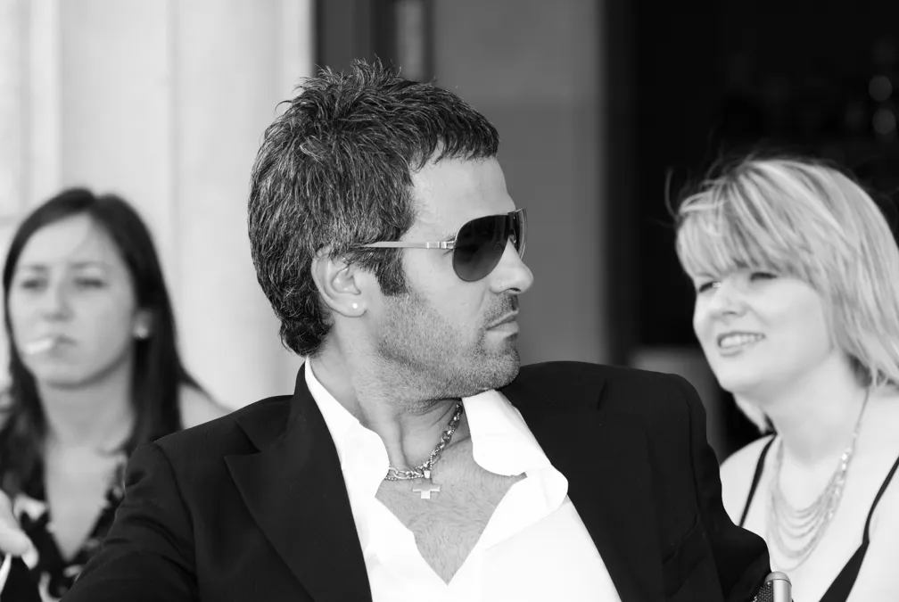 Black and white image of a man in sunglasses and a suit looking to the side with two women in the background, one smiling and one smoking.