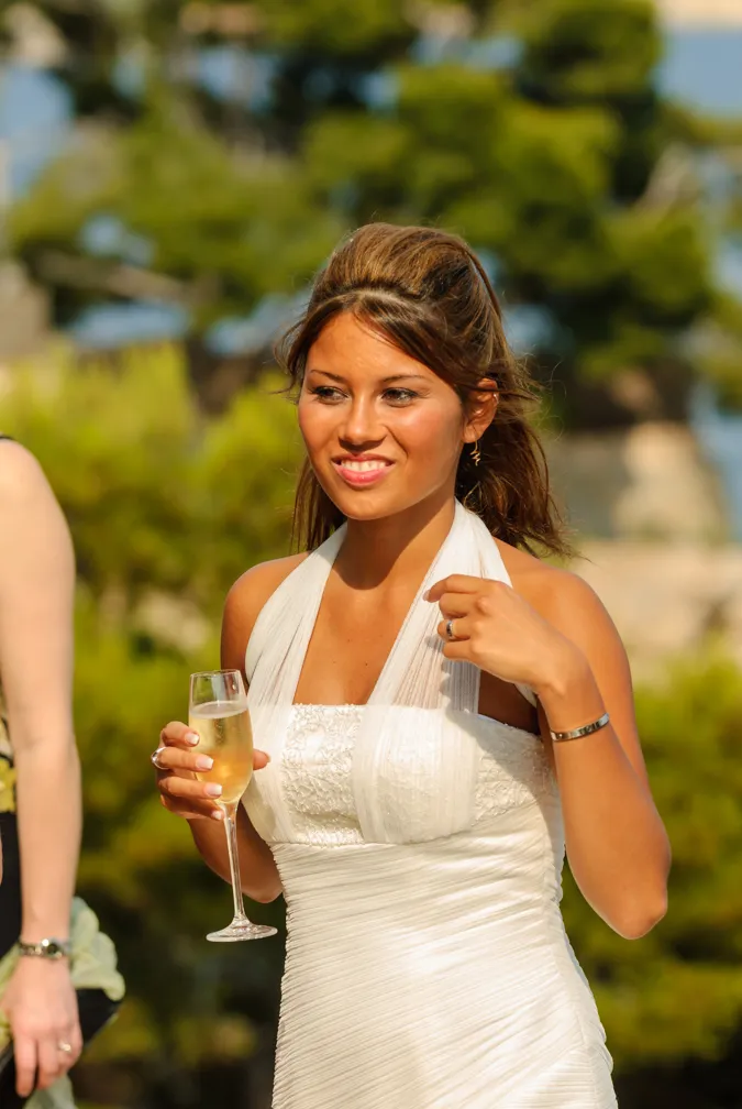 Smiling woman in a white dress holding a glass of champagne outdoors with greenery in the background.