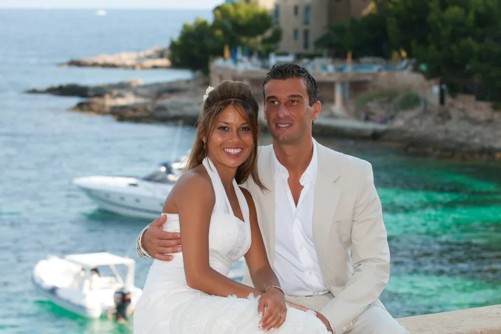 Smiling bride in a white dress and groom in a light beige suit posing by the sea with boats and rocky coastline in the background.