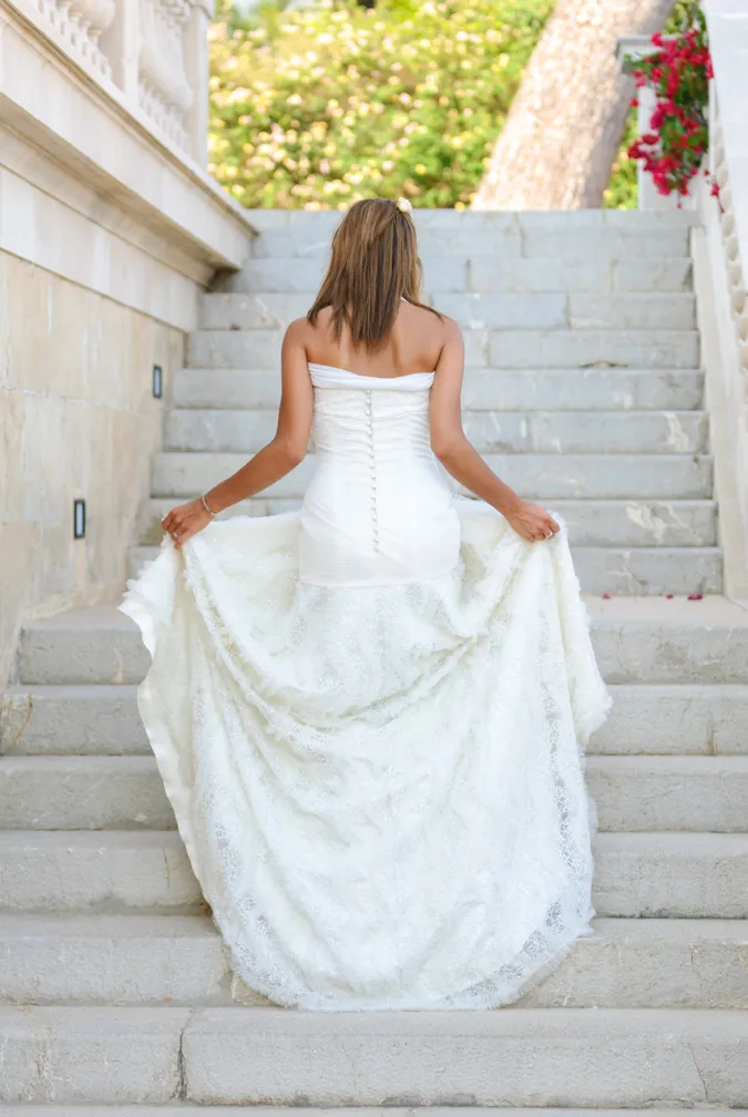 Woman in a strapless white lace wedding dress holding the skirt while ascending stone stairs outdoors.