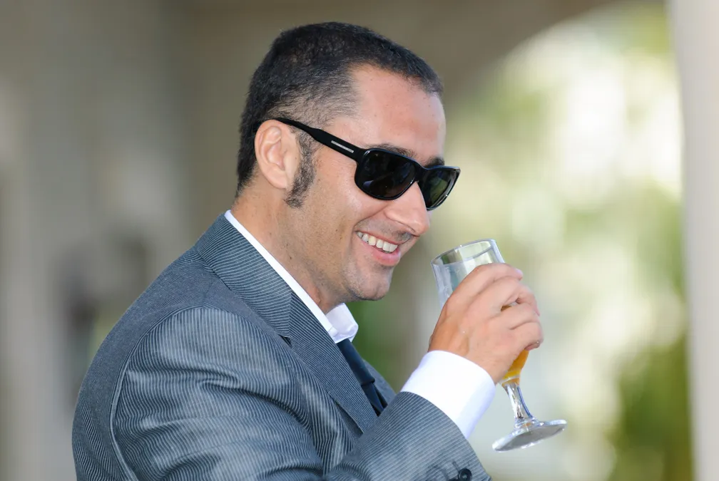 Smiling man wearing sunglasses and a suit holding a glass with amber liquid outdoors.