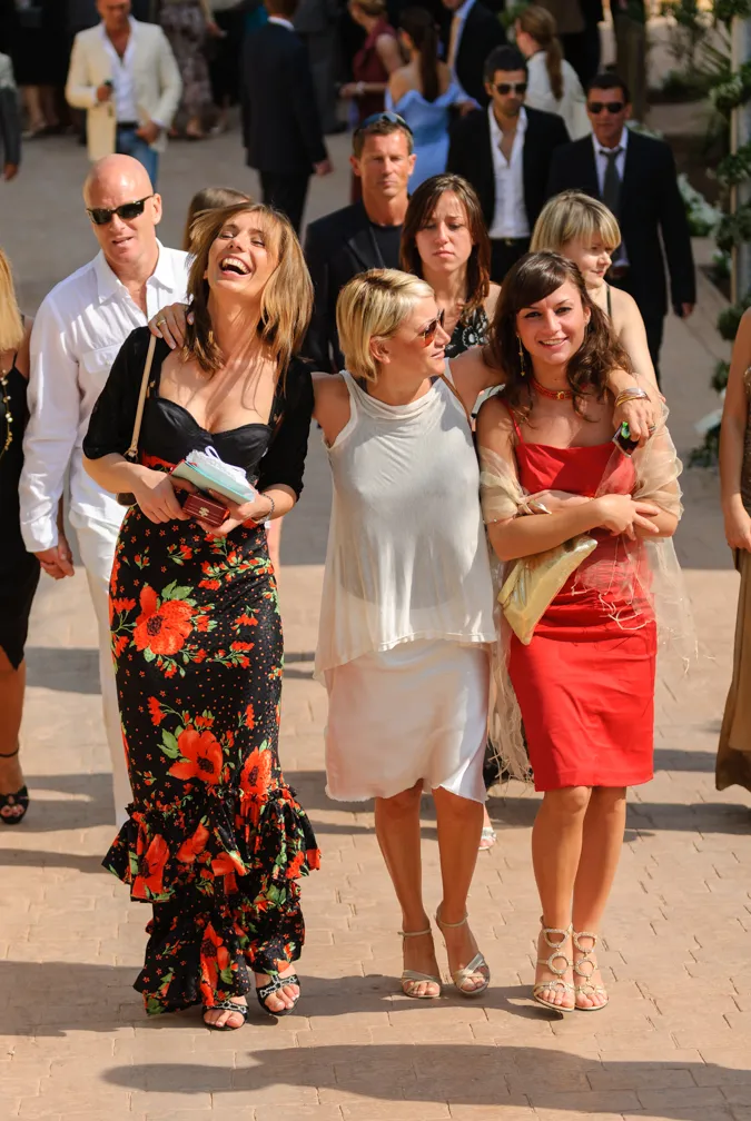Three women walking arm-in-arm outdoors with two wearing dresses and one in a sleeveless top and skirt, smiling and enjoying sunny weather.