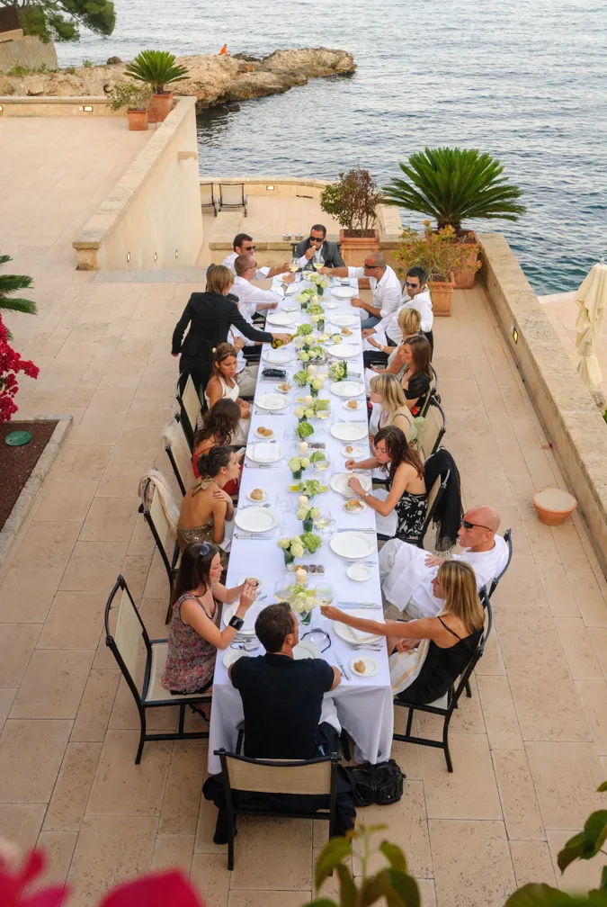 Group of people seated around a long white-clothed table with floral centerpieces dining outdoors near a stone terrace and ocean.
