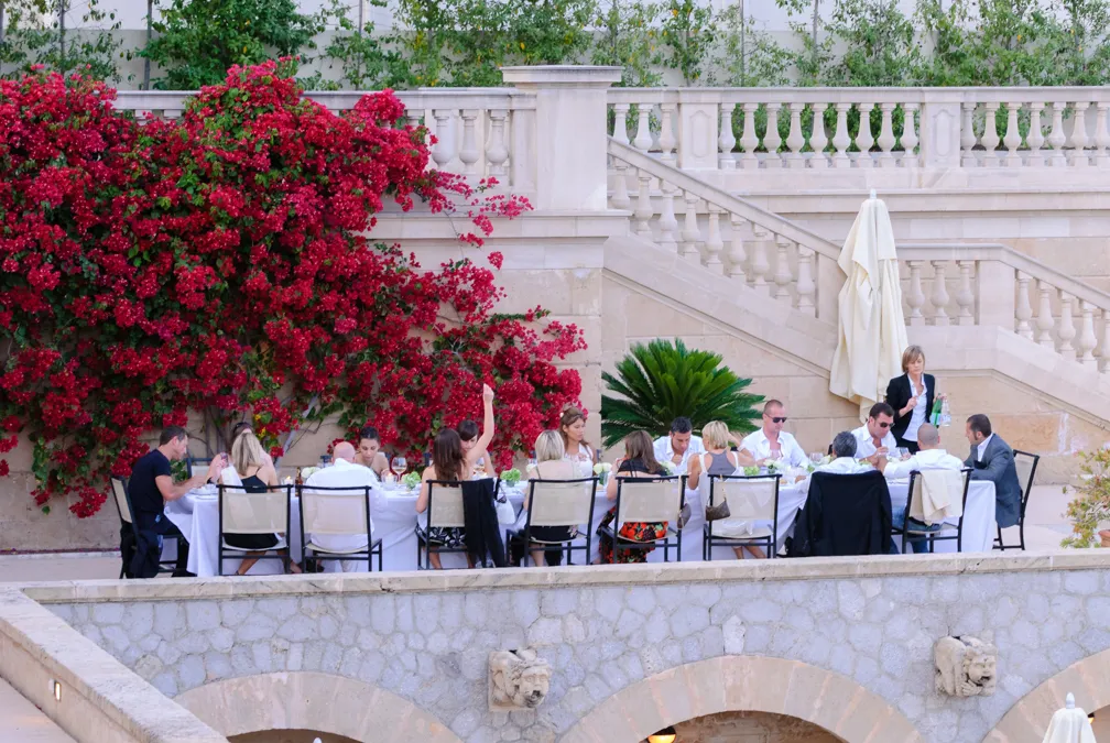 Group of people seated at a long outdoor dining table with white tablecloth, near stone stairs and vibrant red flowering bush.