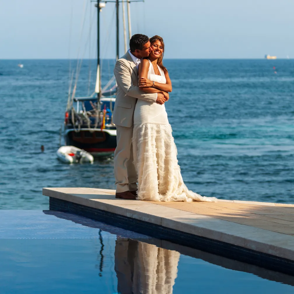 Bride and groom embracing on a pier by the ocean with a sailboat in the background.