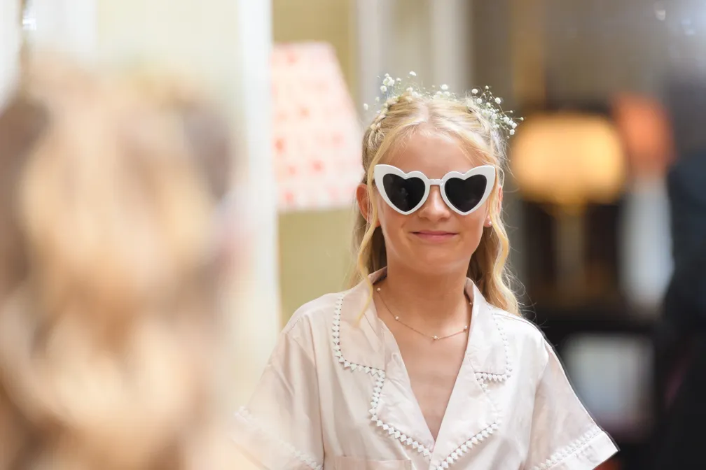 Smiling young woman wearing white heart-shaped sunglasses and a white blouse with floral hair accessories.