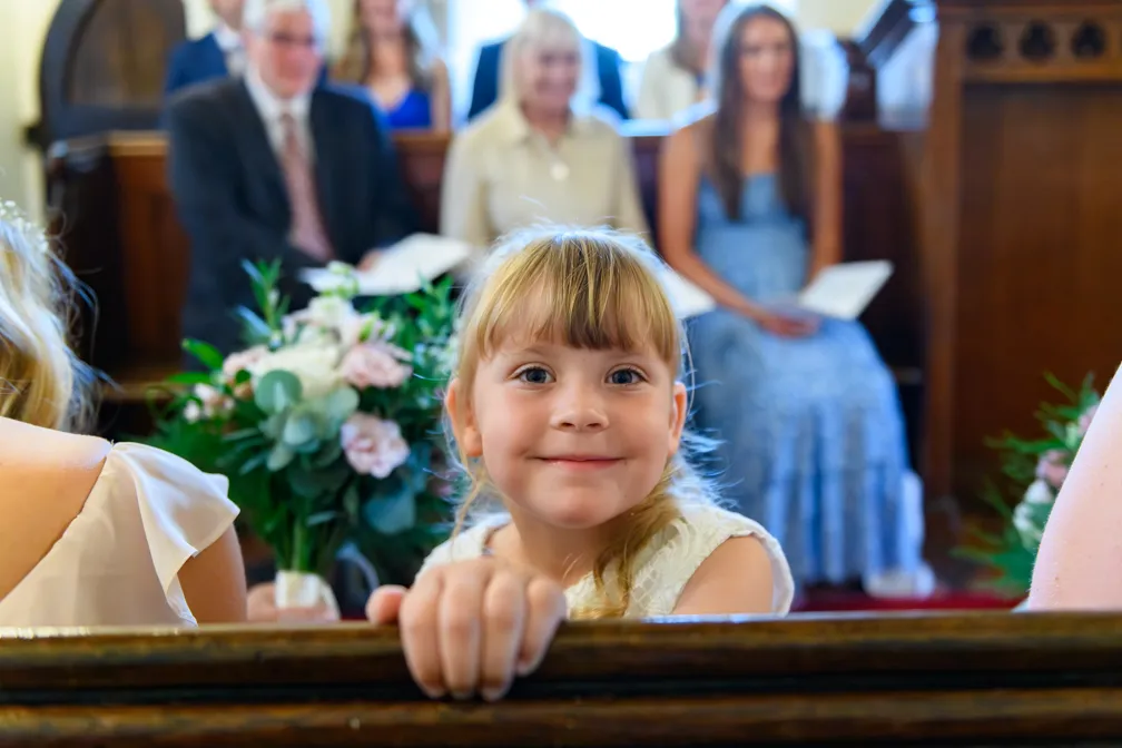 Smiling young girl in a white dress leaning on a wooden pew inside a church, with a floral bouquet and blurred guests in the background.