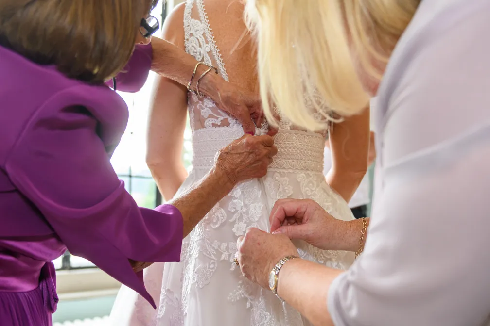 Two women helping to button up the back of a bride's lace wedding dress.