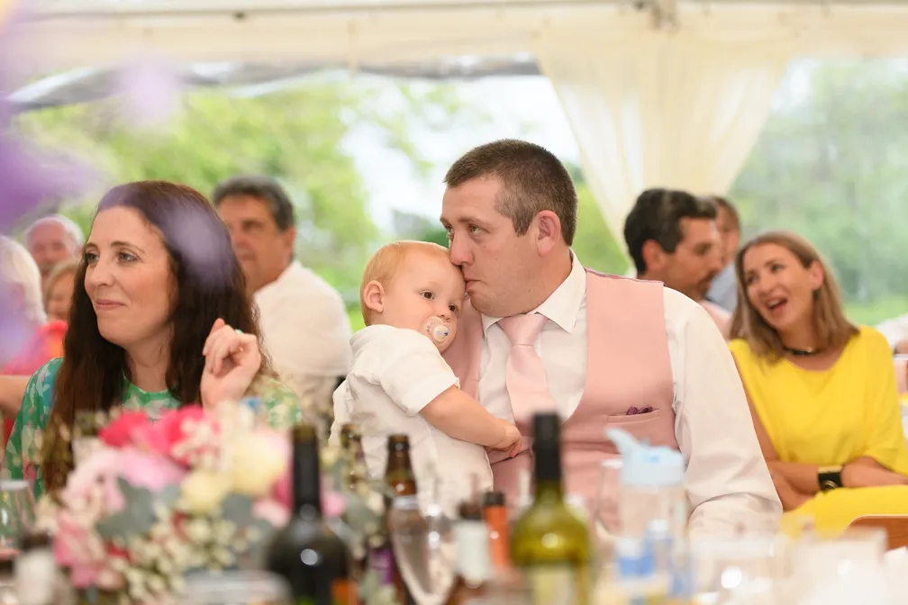 Man in a pink vest and tie holding a baby with a pacifier, seated at a table with a woman in a green floral dress and other guests in the background.