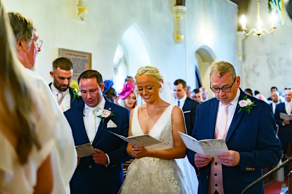 Bride and groom standing at the altar in a church, reading from ceremony programs, surrounded by guests.