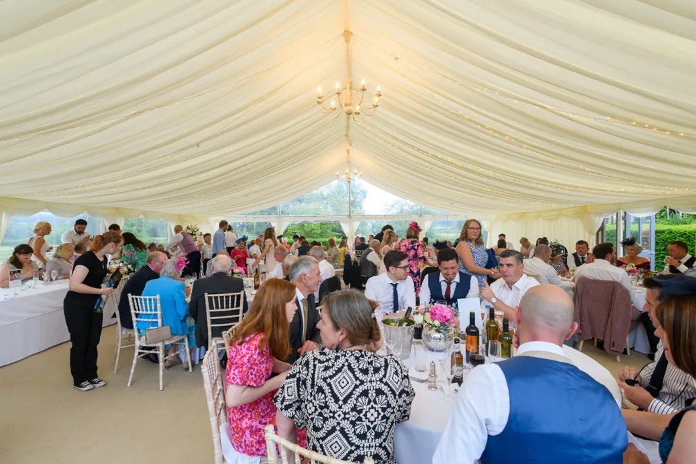 Guests seated at decorated tables inside a white marquee with draped ceiling and chandeliers, attending a formal event.