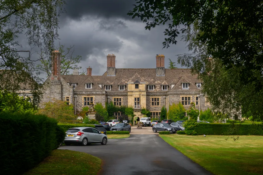 Large historic stone mansion with multiple chimneys and parked cars on a driveway under a cloudy sky.