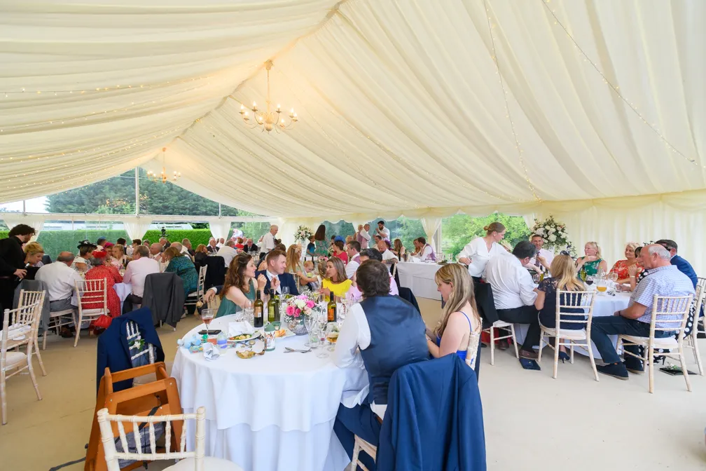 Guests seated at round tables under a white tent with draped ceiling and string lights at a formal event.
