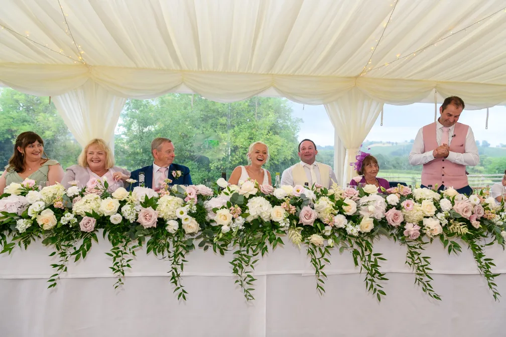 Wedding party seated at a long table adorned with white and pink floral arrangements under a white tent.