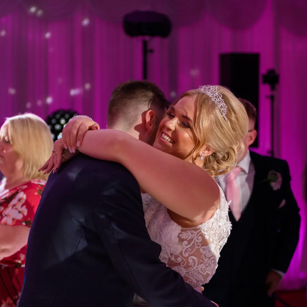 Bride smiling and hugging groom during wedding celebration with purple-lit background.