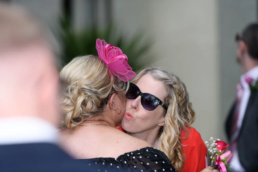 Woman wearing sunglasses and red outfit giving a cheek kiss to another woman with blonde hair and a pink fascinator.