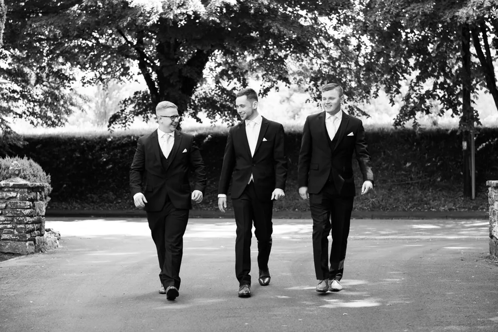 Three men in suits walking and smiling outdoors under large trees.