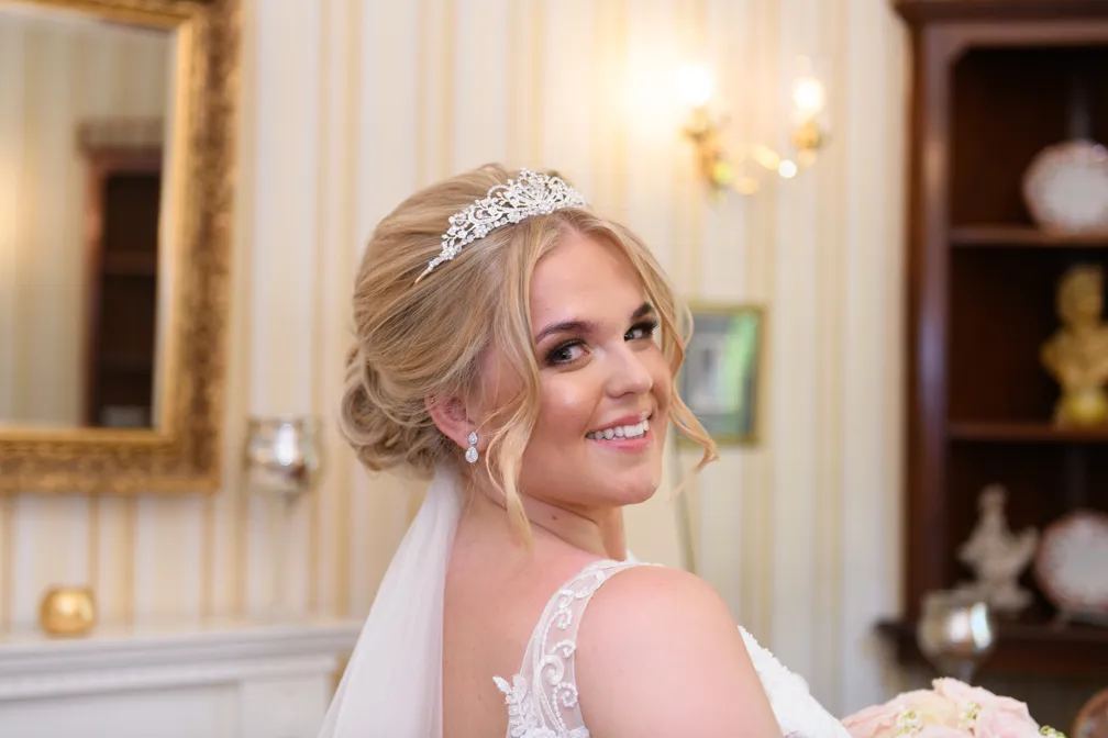 Smiling bride with blonde hair in an updo wearing a tiara and lace wedding dress in an elegant room.