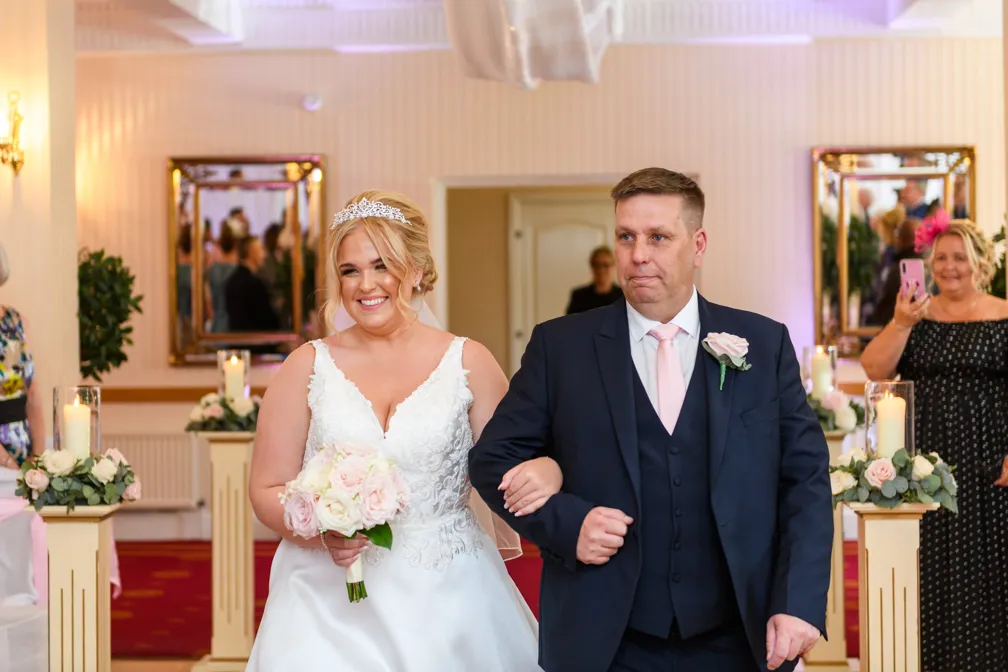 Bride in white gown and tiara holding bouquet, walking arm-in-arm with man in navy suit and pink tie at wedding ceremony.