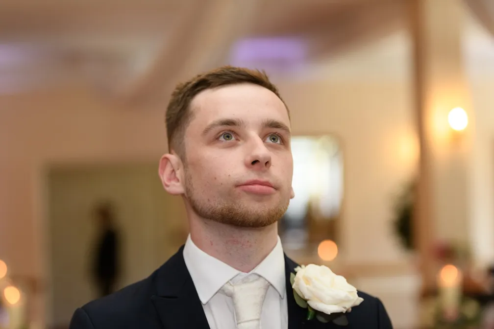Young man in a suit with a white rose boutonniere looking slightly upward indoors.