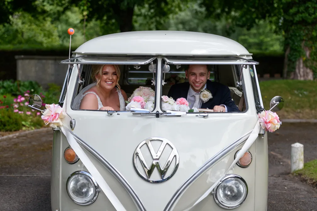 Smiling bride and groom sitting in a decorated white vintage Volkswagen van with floral ribbons on side mirrors.