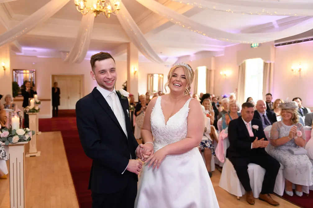 Smiling bride in a white wedding dress and groom in a black suit holding hands in a decorated wedding venue with seated guests clapping.