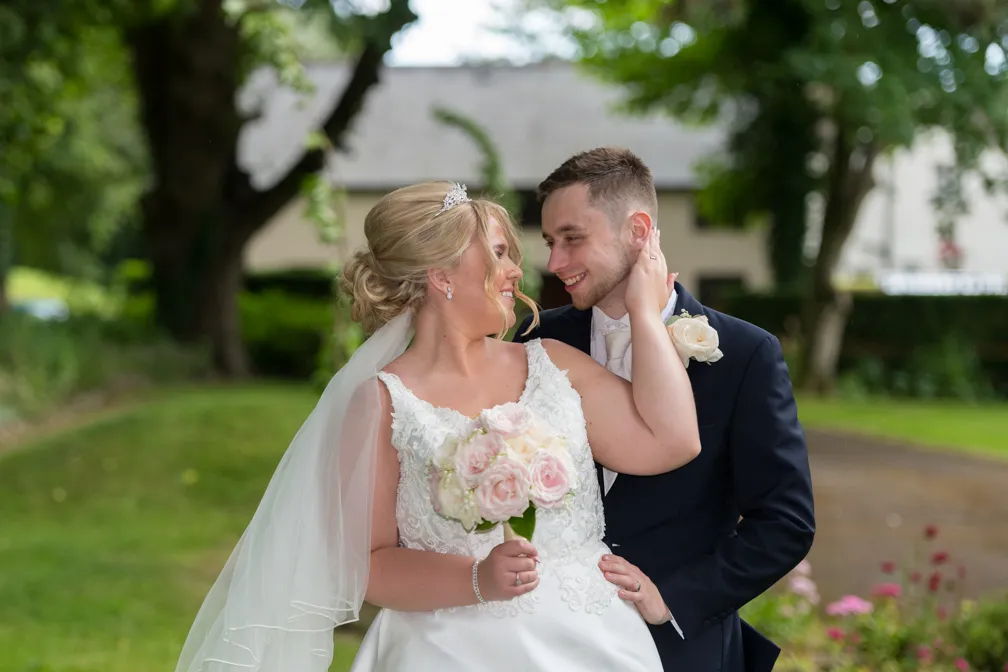 Bride in white dress holding pink and white bouquet smiling at groom in black suit with white rose boutonniere outdoors.