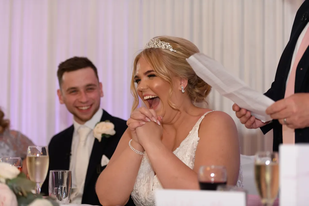 Bride laughing joyfully during a wedding speech with groom smiling in the background.
