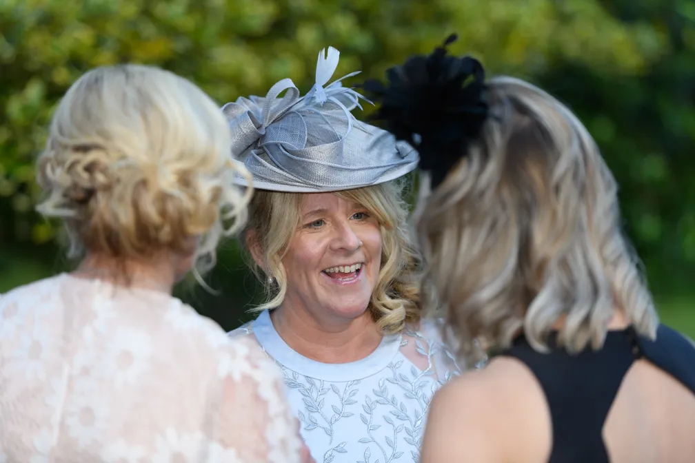 Smiling woman in a light blue dress with floral embroidery wearing a grey fascinator, talking to two women with braided hairstyles.