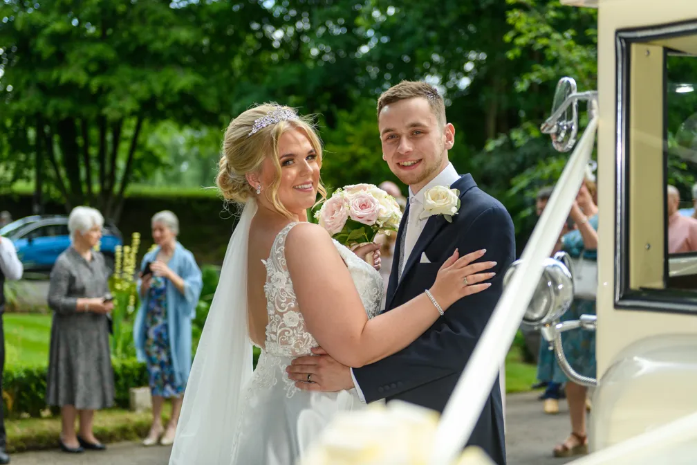 Bride and groom smiling and embracing outdoors near a classic white car with green trees and guests in the background.