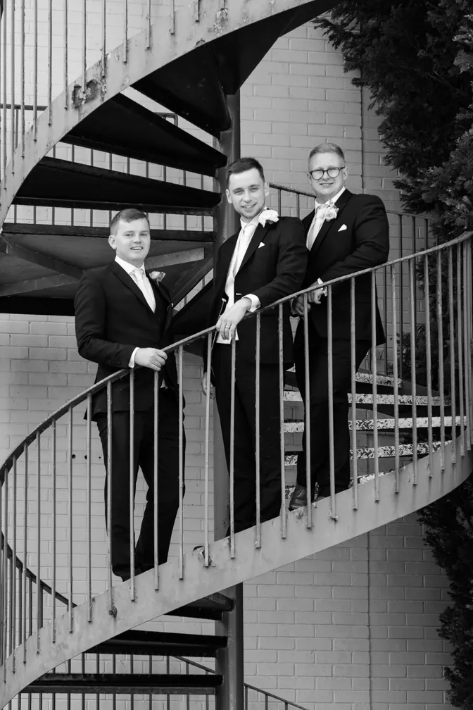Three men in tuxedos standing on a spiral staircase against a brick wall, smiling at the camera in a black and white photo.